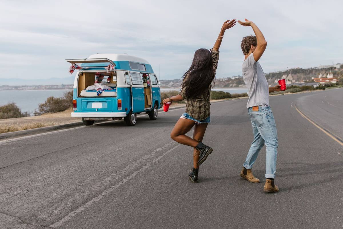Two backpackers dancing on a coastal road next to their camper van