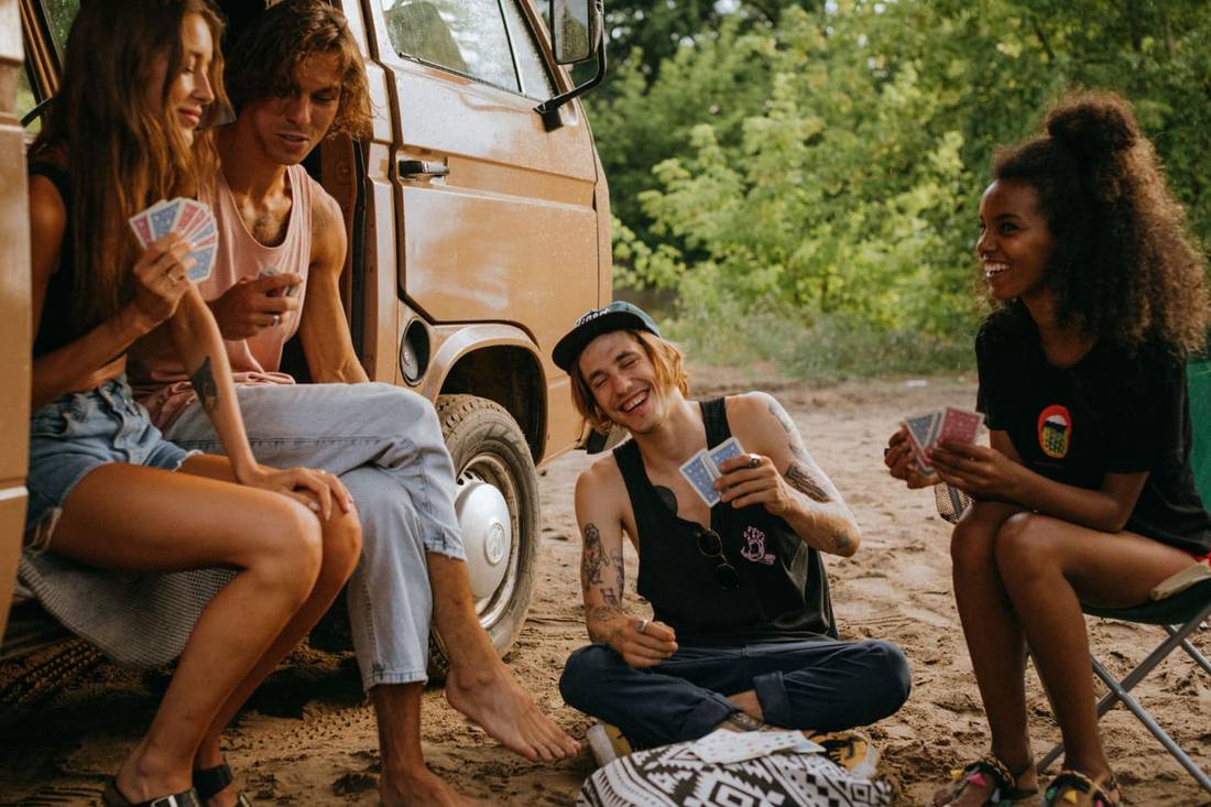 Group of friends playing cards by a camper van in a sunny outdoor setting.