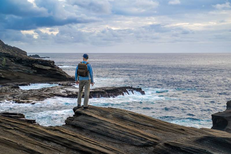 A man stands on a rocky cliff overlooking the ocean, with a backpack, under a cloudy sky.